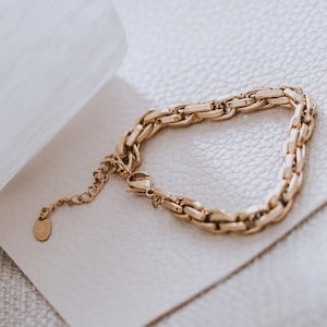 May include: A gold chain bracelet with a chunky, textured design. The bracelet has a small, round tag attached to it.
