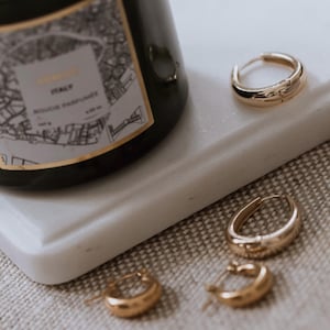 May include: Three gold hoop earrings of varying sizes on a white marble surface. The earrings are thick and have a slightly hammered texture.