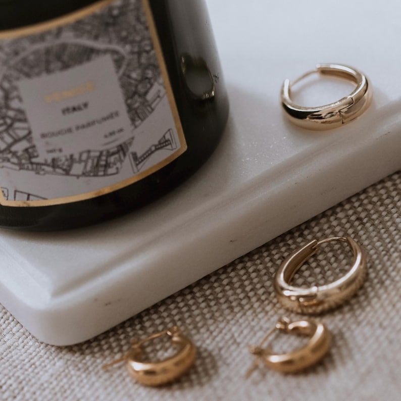 May include: Three gold hoop earrings of varying sizes on a white marble surface. The earrings are thick and have a slightly hammered texture.