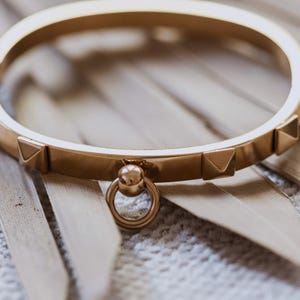 Puede incluir: Un brazalete de oro con un peque&ntilde;o dije redondo de oro adjunto. El brazalete tiene una serie de peque&ntilde;os tachuelas triangulares a lo largo del borde.