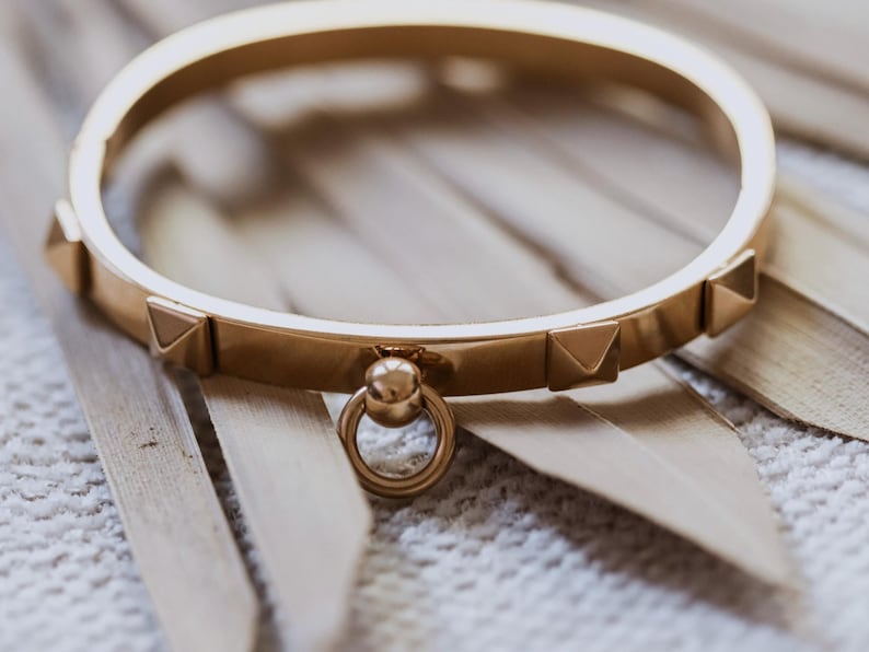 Puede incluir: Un brazalete de oro con un peque&ntilde;o dije redondo de oro adjunto. El brazalete tiene una serie de peque&ntilde;os tachuelas triangulares a lo largo del borde.