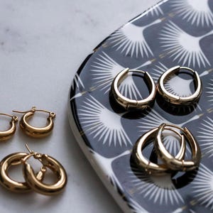 May include: Gold hoop earrings of different sizes on a black and white patterned tray. The earrings are all oval shaped and have a smooth finish.