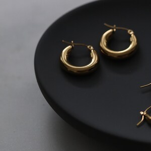 May include: Three pairs of gold hoop earrings of different sizes on a black plate. The earrings are all smooth and have a simple design.