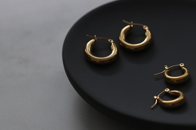May include: Three pairs of gold hoop earrings of different sizes on a black plate. The earrings are all smooth and have a simple design.