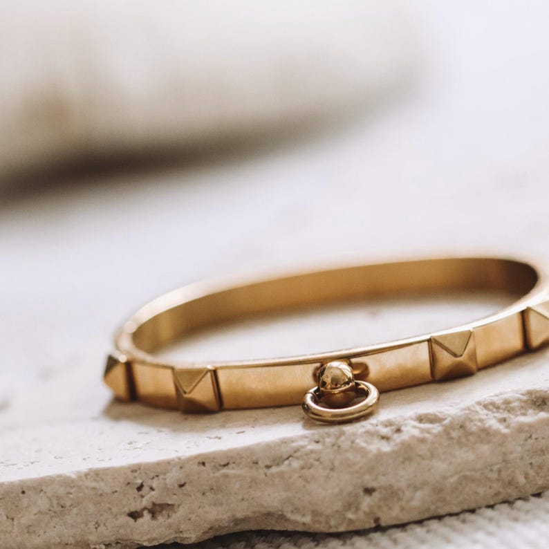 Puede incluir: Un brazalete dorado con un peque&ntilde;o charm redondo colgante. El brazalete tiene una serie de peque&ntilde;os acentos en forma de pir&aacute;mide en la parte superior.
