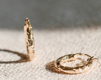 May include: Two gold hoop earrings with a textured, irregular surface. The earrings are on a light beige fabric background.
