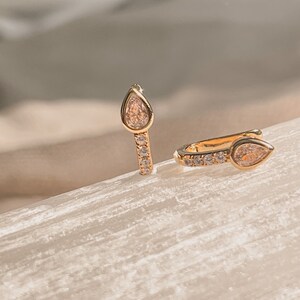 May include: A pair of gold-coloured earrings. Each earring has a pear-shaped, peach-coloured gemstone at the top. The earring's body is decorated with small, clear gemstones. The earrings are displayed on a light-coloured surface.
