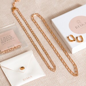 Puede incluir: Un conjunto de collares de cadena dorada y pendientes de aro dorados en una caja blanca con un logotipo circular rosa que dice "RIAKA Jewelry". Los collares est&aacute;n en una bolsa de pl&aacute;stico transparente con una tarjeta rosa que dice "Tengo suficientes joyas, nunca lo dijo nadie."