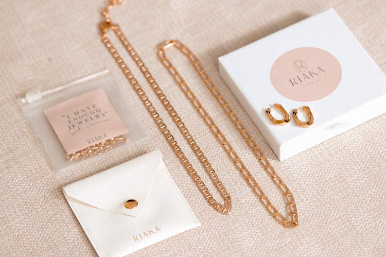 Puede incluir: Un conjunto de collares de cadena dorada y pendientes de aro dorados en una caja blanca con un logotipo circular rosa que dice "RIAKA Jewelry". Los collares est&aacute;n en una bolsa de pl&aacute;stico transparente con una tarjeta rosa que dice "Tengo suficientes joyas, nunca lo dijo nadie."