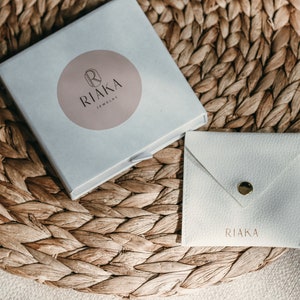 Puede incluir: Una caja blanca con un logotipo circular rosa que dice "RIAKA Jewelry". La caja est&aacute; sentada sobre una estera de paja tejida. Tambi&eacute;n hay un sobre blanco con la palabra "RIAKA" impresa en &eacute;l.