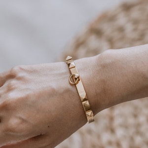 Puede incluir: Un brazalete dorado con un cierre decorativo y una fila de tachuelas en forma de pir&aacute;mide.