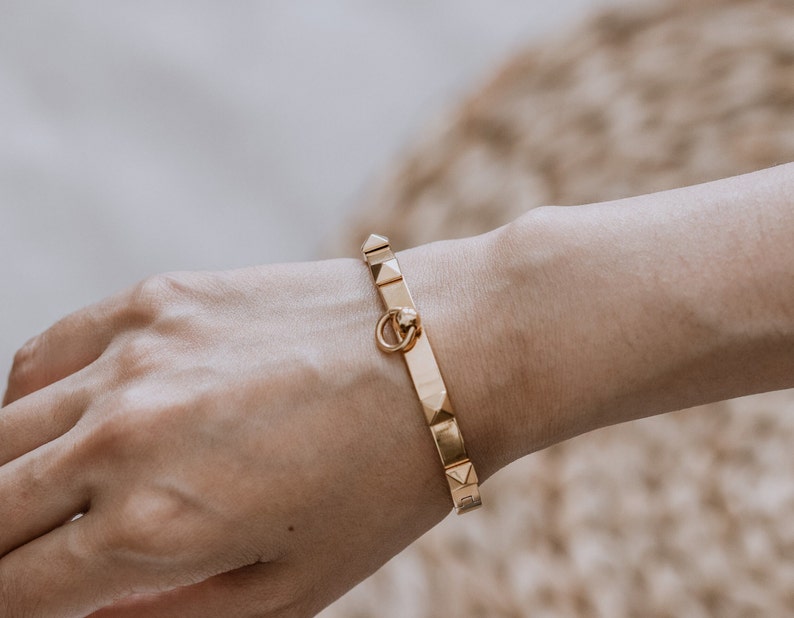 Puede incluir: Un brazalete dorado con un cierre decorativo y una fila de tachuelas en forma de pir&aacute;mide.