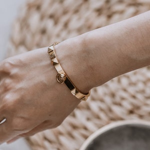 Puede incluir: Un brazalete dorado con un dise&ntilde;o texturizado y tachonado. El brazalete tiene un peque&ntilde;o cierre redondo.