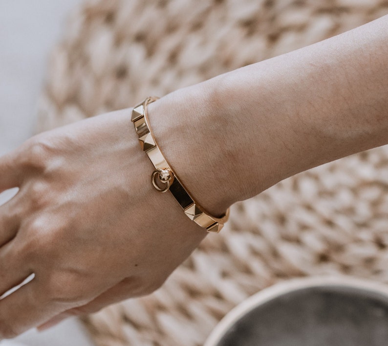 Puede incluir: Un brazalete dorado con un dise&ntilde;o texturizado y tachonado. El brazalete tiene un peque&ntilde;o cierre redondo.