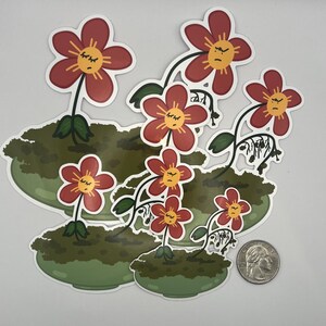Puede incluir: Un conjunto de pegatinas con flores de dibujos animados con pétalos rojos y centros amarillos, cada una con una expresión de mal humor. Las flores están en macetas verdes con aspecto de musgo. Se incluye una moneda como referencia de tamaño.