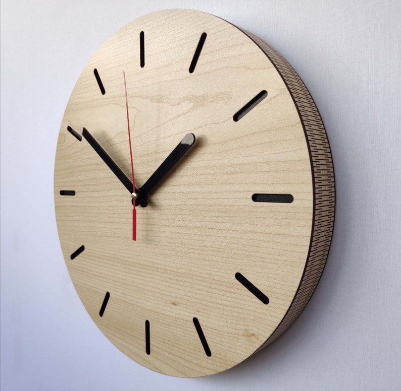May include: A round wooden clock with black hour and minute hands and black markings for the hours. The clock face is light wood with a darker wood grain pattern.