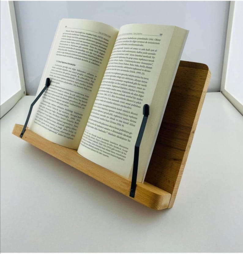 Pode incluir: Um suporte de livro de madeira com suportes de p&aacute;gina de metal preto. O suporte &eacute; feito de madeira marrom claro e tem um acabamento natural. O suporte est&aacute; segurando um livro aberto com texto em um idioma estrangeiro.