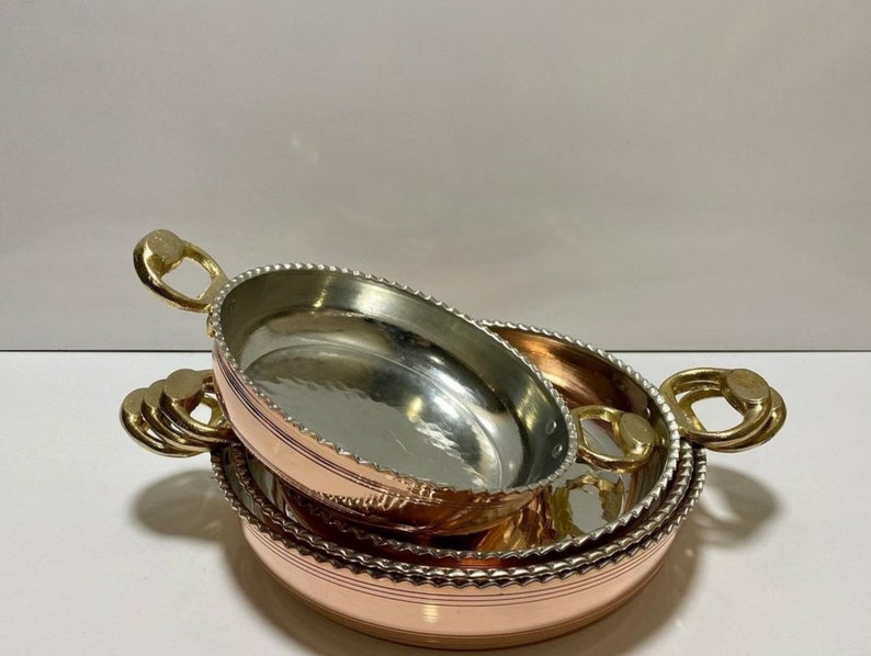 K&ouml;nnte beinhalten: Ein Satz aus drei Kupfer-Servierschalen mit Messinggriffen. Die Schalen sind ineinander verschachtelt, wobei die gr&ouml;&szlig;te Schale unten und die kleinste oben liegt. Die Schalen haben eine geh&auml;mmerte Oberfl&auml;che und einen perlenbesetzten Rand.