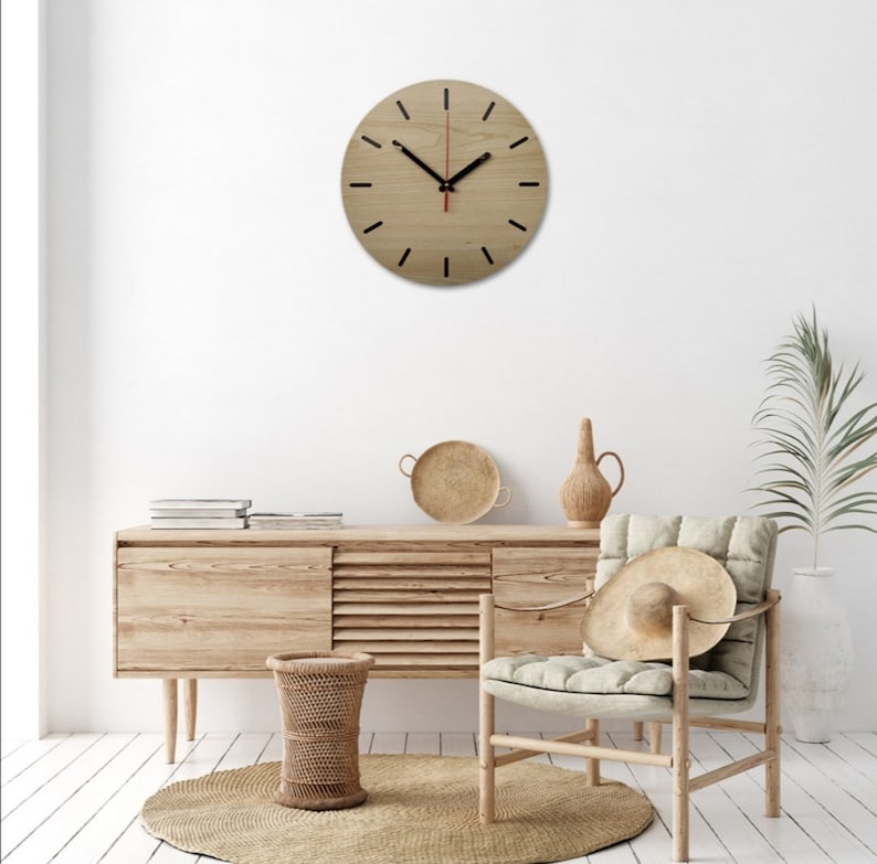 May include: A wooden wall clock with black hour markers and a red second hand. The clock is hanging on a white wall above a wooden dresser with a woven basket and a chair with a straw hat.