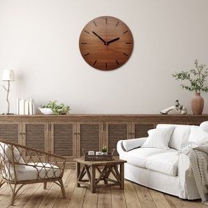 May include: A brown wooden wall clock with black hour markers and a red second hand. The clock is hanging on a white wall in a living room setting with a couch, coffee table, and plants.