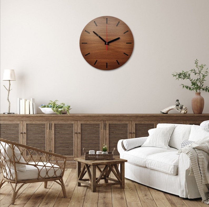 May include: A brown wooden wall clock with black hour markers and a red second hand. The clock is hanging on a white wall in a living room setting with a couch, coffee table, and plants.