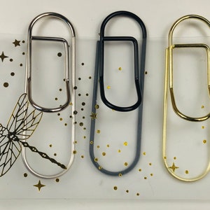 May include: Three binder clips in silver, black, and gold. The clips are on a clear background with a gold dragonfly and gold stars.