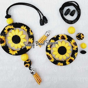 May include: A set of sunflower-themed jewelry supplies, including a necklace with a large sunflower charm, a smaller sunflower charm, a tassel, and a set of yellow and black beads. The necklace is made with black elastic cord and has a black toggle clasp. The charms are made of polymer clay and have a glossy finish. The beads are also made of polymer clay and have a matte finish. The set is perfect for making your own sunflower-themed jewelry.