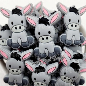 May include: A group of gray donkey silicone charms with pink ears and black eyes. The charms are in a pile and some are partially obscured by others.