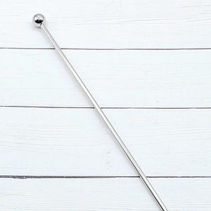 May include: A stainless steel cocktail muddler with a spherical top and a flat, paddle-shaped end. The long, slender tool is designed for mixing drinks. The muddler is shown against a white wooden background.