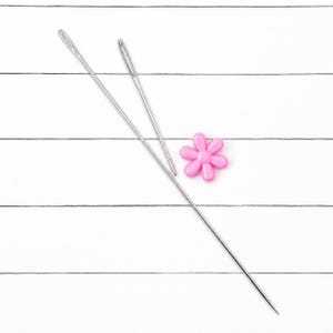 May include: Two long, slender silver metal sewing needles with large eyes positioned diagonally across the frame, accompanied by a small, six-petaled pink flower-shaped plastic button or embellishment resting between the needles.