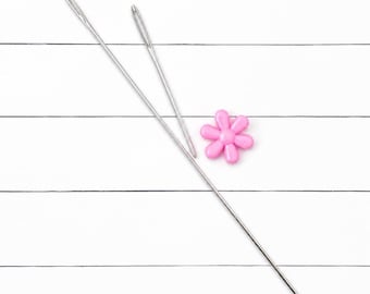 May include: Two long, slender silver metal sewing needles with large eyes positioned diagonally across the frame, accompanied by a small, six-petaled pink flower-shaped plastic button or embellishment resting between the needles.