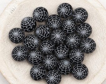 May include: A collection of round black beads, each featuring a white printed spider web pattern. The beads are spherical in shape and display a consistent graphic design across their surfaces, arranged in a cluster on a textured surface.