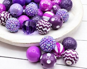 May include: A collection of round beads in various shades of purple, including solid, glittery, translucent, and faceted finishes. Some beads feature white chevron patterns, white polka dots, or white stripes, all with central holes for stringing.
