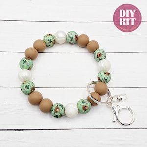May include: A beaded keychain bracelet with a silver clasp. The bracelet features a mix of brown, white, and green beads. The green beads have a pattern of a brown animal and green leaves. The text "DIY KIT EVERYTHING INCLUDED" is in a pink circle.