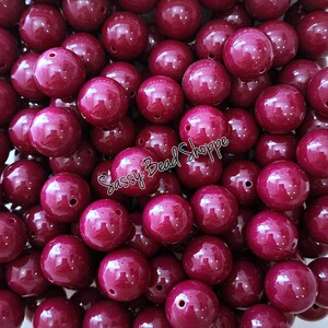 May include: A collection of glossy, round, burgundy beads with drilled holes. The beads are scattered, filling the frame. The text "Sassy Bead Shoppe" is visible in the image. These beads are suitable for jewelry making and crafts.