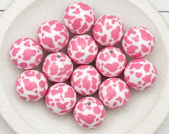 May include: A collection of twelve spherical beads featuring a white base with a pink cow print pattern. Each bead has a small, centered hole for threading, and they are arranged in a cluster on a light-colored, circular wooden surface.