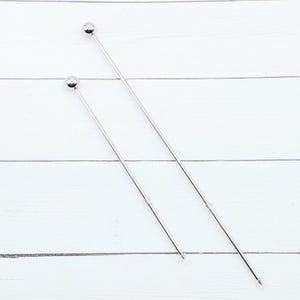 May include: Two silver cocktail stirrers with a ball at the top and a pointed end. The stirrers are positioned diagonally against a white wooden background. These bar accessories are ideal for mixing drinks.