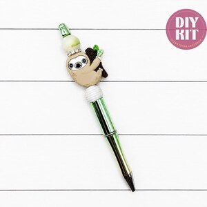 May include: A metallic green pen featuring a decorative topper with a tan sloth character clinging to a brown branch, accented by a white and green marbled bead, a silver rhinestone spacer, and a white ribbed bead. The text DIY KIT is visible.