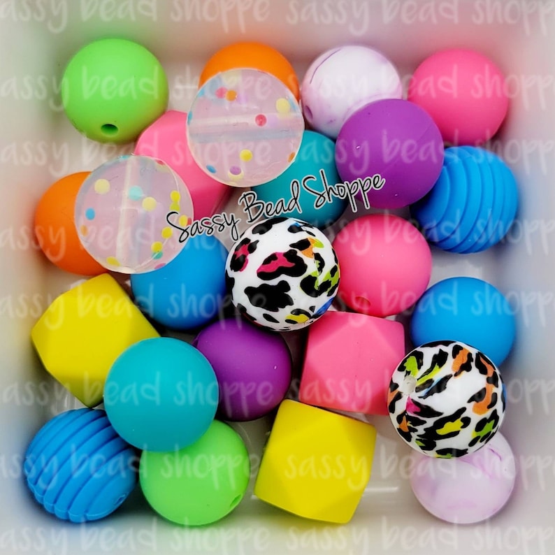 K&ouml;nnte beinhalten: Eine Sammlung bunter, runder und quadratischer Silikonperlen in verschiedenen Mustern, darunter Leopardenmuster, Punkte und Streifen. Die Perlen sind in einer wei&szlig;en Sch&uuml;ssel angeordnet. Der Text "Sassy Bead Shoppe" ist in der Mitte des Bildes sichtbar.
