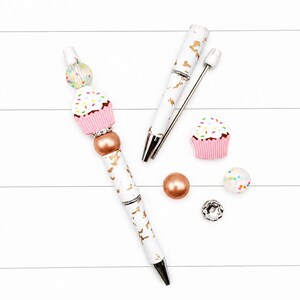 Op de afbeelding: Een witte balpen met goudfolie accenten, voorzien van een siliconen cupcake kraal, een doorzichtige kraal met gekleurde stippen, een koperkleurige metalen kraal en een strass tussenstuk. Inclusief een bijpassende penhuls en losse kralen.