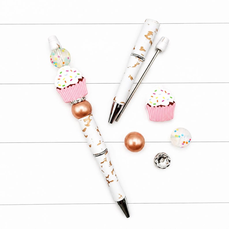Op de afbeelding: Een witte balpen met goudfolie accenten, voorzien van een siliconen cupcake kraal, een doorzichtige kraal met gekleurde stippen, een koperkleurige metalen kraal en een strass tussenstuk. Inclusief een bijpassende penhuls en losse kralen.
