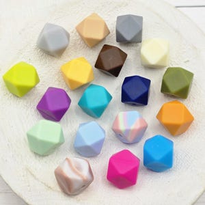 May include: A collection of colorful, geometric silicone beads in various shades including gray, yellow, brown, blue, green, purple, pink, and orange. The beads are arranged on a white surface, showcasing their multifaceted design.