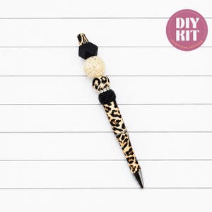 May include: A beaded pen featuring a leopard print pattern on the barrel and upper beads, accented by a black geometric bead, a textured gold bead, and a black ribbed bead with a silver-toned ring. The text DIY KIT is visible in a circle.