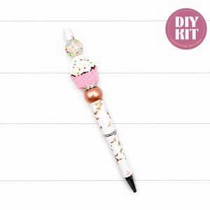 Op de afbeelding: Een witte balpen met een goud gespikkelde houder en een decoratieve topper bestaande uit een doorzichtige kraal met gekleurde stippen, een roze cupcake-bedel met wit glazuur en hagelslag, en een koperkleurige kraal met zilveren accenten. Tekst: DIY KIT.