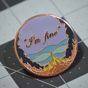 May include: A round enamel pin with a purple and blue cloud with the text "I'm fine" in white. The cloud is over a yellow and orange flame design.