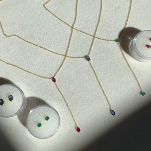 May include: Three gold necklaces with different colored gemstones at the end. The necklaces are layered on a white surface. The gemstones are red, blue, and green.