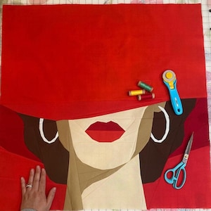 Puede incluir: Un bloque de colcha de tela roja con la cara de una mujer en el centro. La cara está hecha de piezas de tela en tonos de marrón, beige y blanco. La mujer tiene labios rojos y grandes pendientes blancos. El bloque de colcha está rodeado de tela roja.
