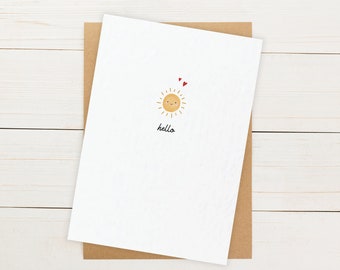 May include: A white greeting card with a smiling sun illustration, two small red hearts, and the word "hello" in black script. The card is on a brown envelope, set against a white wooden background.