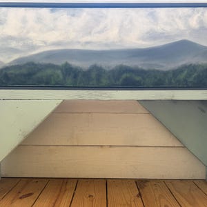 May include: A framed landscape painting depicting a mountain range with a forest in the foreground. The artwork is displayed on a light green wooden bench with angled legs. The painting features shades of gray, green, and blue.