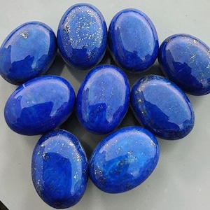 May include: Nine oval-shaped, polished lapis lazuli stones. The stones are a deep, rich blue with flecks of gold, arranged on a light gray surface. The gemstones are smooth and have a glossy finish, reflecting light.